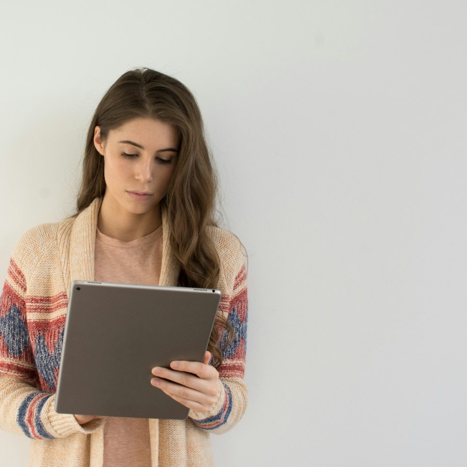Woman in a light cardigan reading from a tablet, standing against a plain wall.
